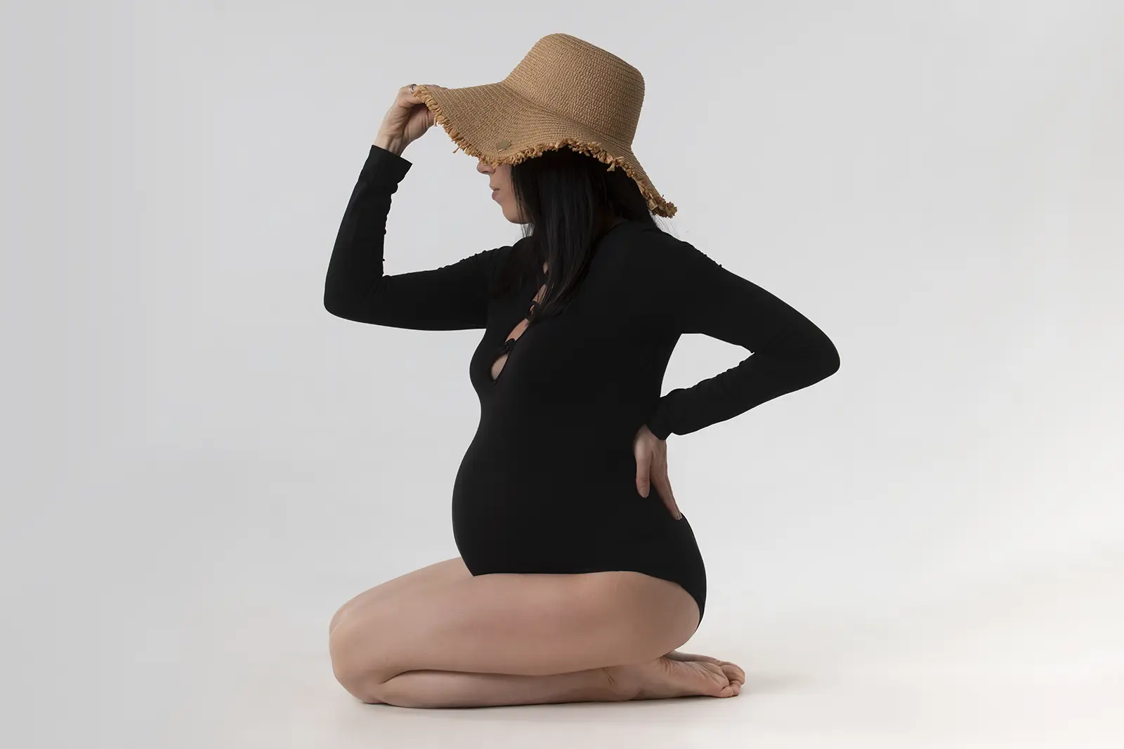 Donna incinta in ginocchio con body nero e cappello di paglia, scatto in studio a Milano
