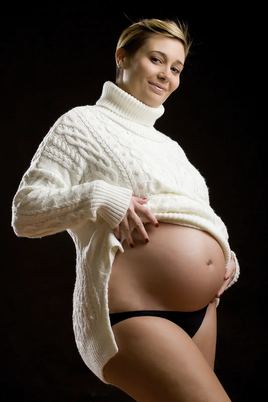 Futura mamma con maglione bianco e pancione scoperto su fondo scuro, book fotografico gravidanza in studio a Milano